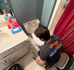 A plumber repairing a toilet in a small bathroom with tools on the floor and a red shower curtain.