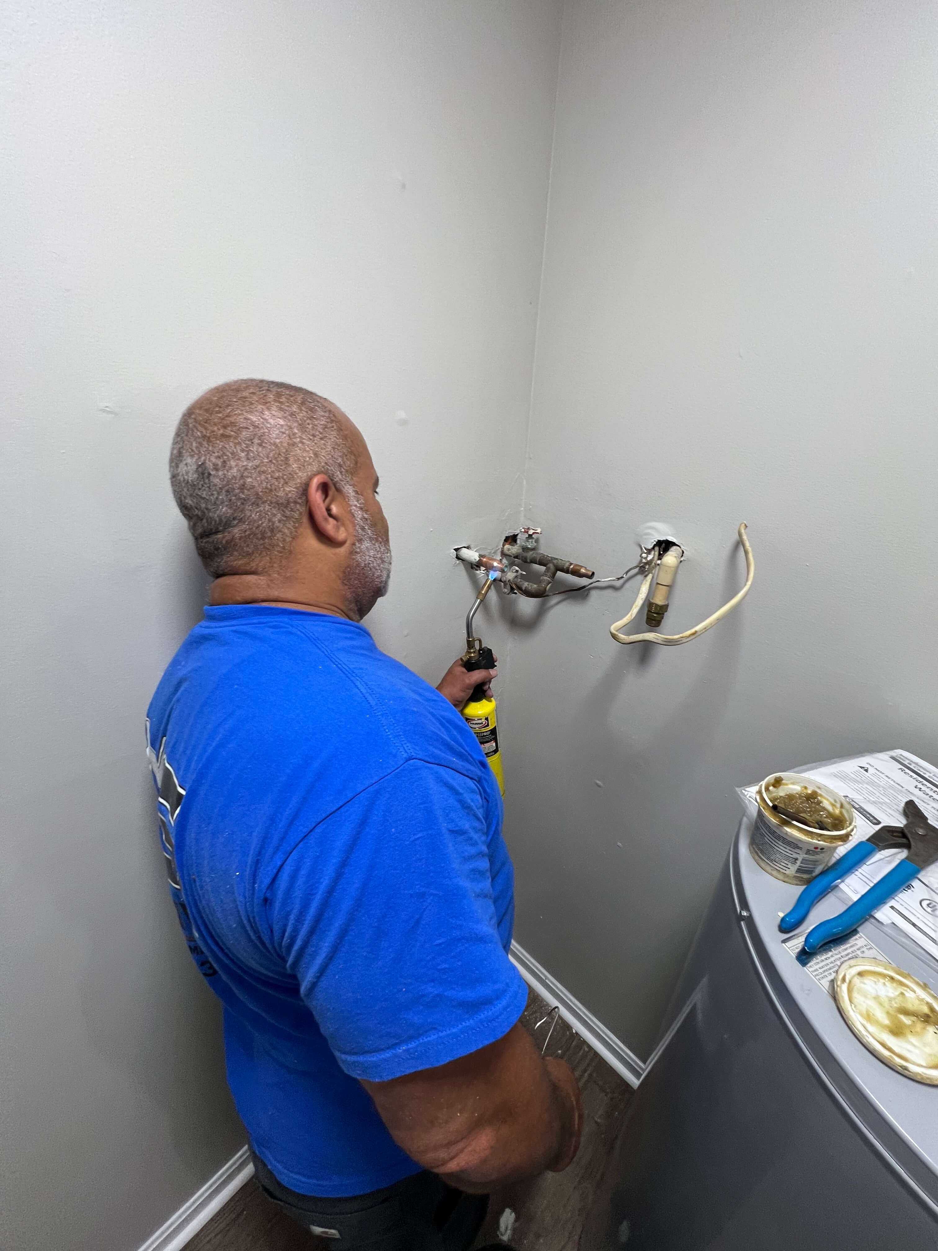 Man in a blue shirt using a torch to solder copper pipes on a gray wall near a water heater with tools and plumbing paste beside it.