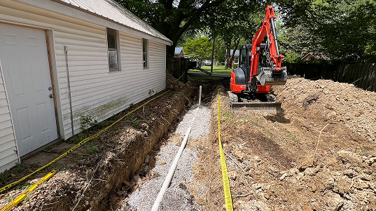 Construction site next to a house with a trench dug for sewer and water pipes, orange excavator and piles of dirt nearby.