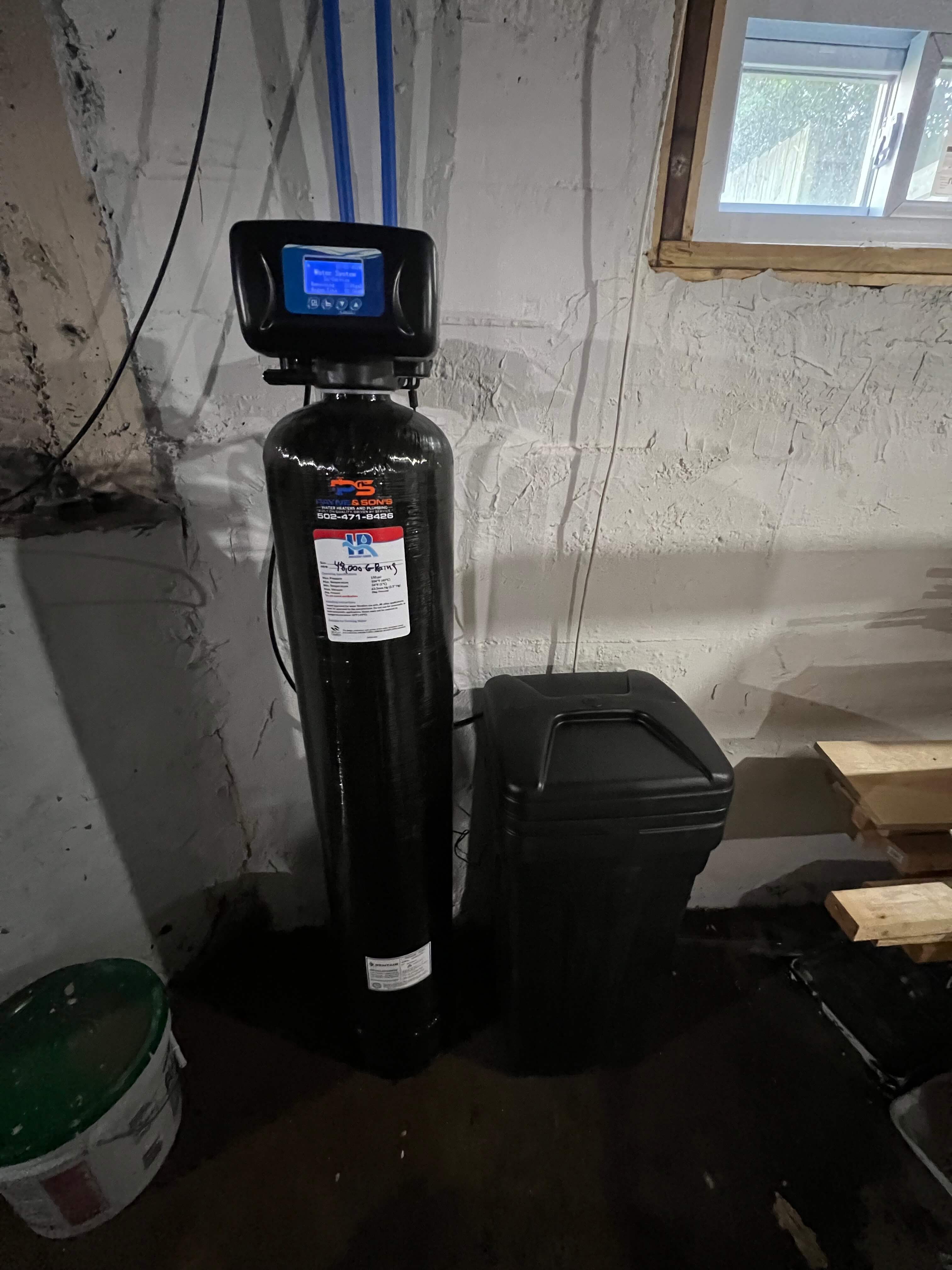Water softener system installed in a basement with a tall black tank and a black brine tank beside it under a small window.