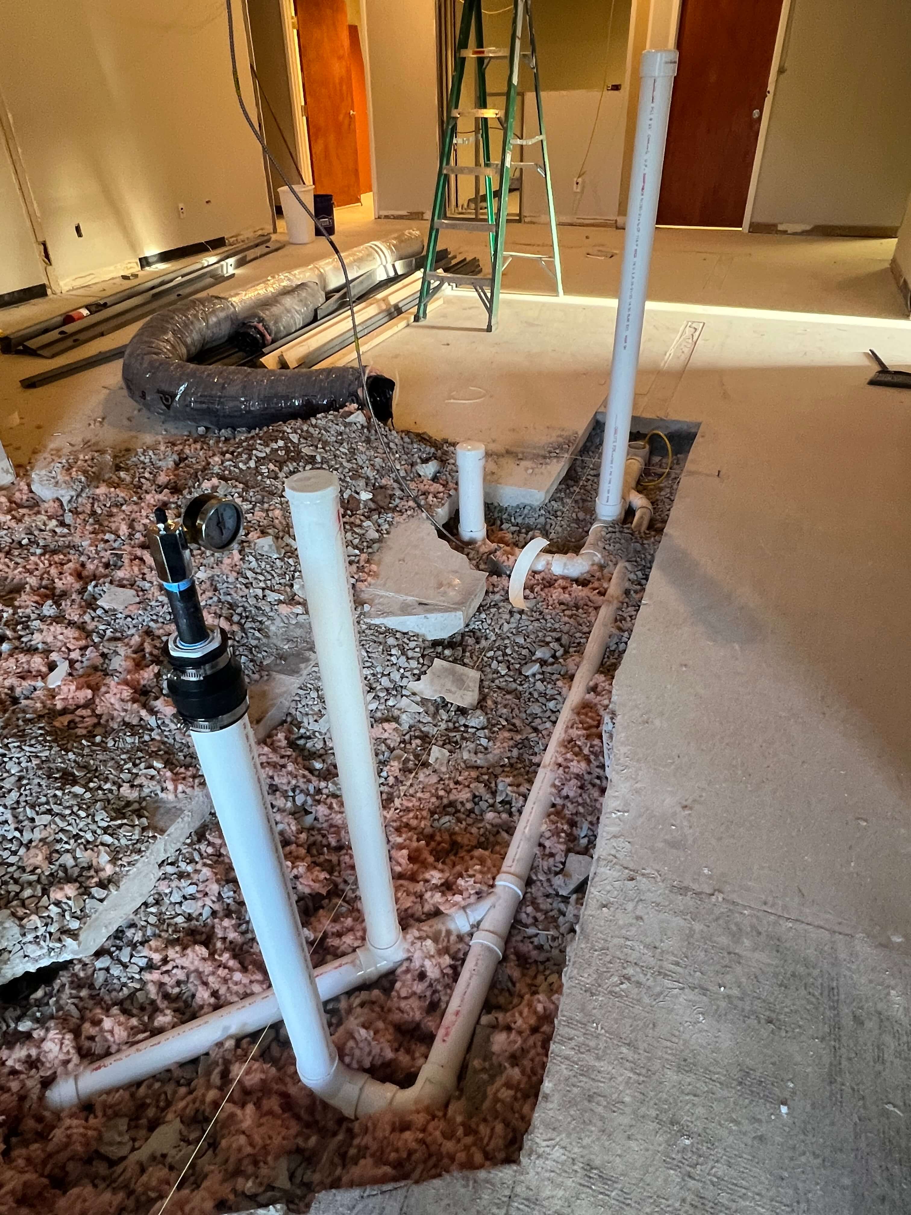 Indoor construction site showing exposed white plumbing pipes installed in a gravel-filled trench beneath a concrete floor.