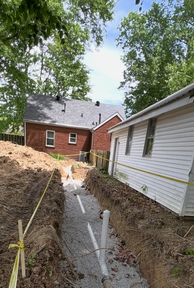 Excavated trench beside a white house with newly installed white plumbing pipes on a bed of gravel, surrounded by trees and a brick house in the background.