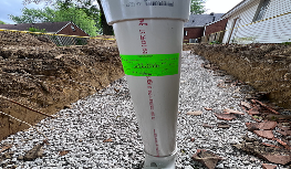 White PVC pipe installed vertically in gravel-filled trench between residential houses for flood drainage.