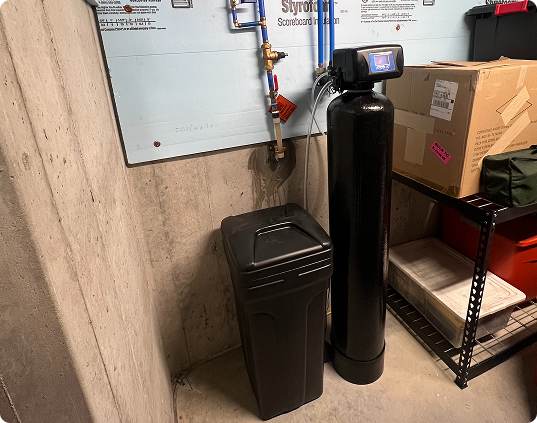 Water filtration system in a basement with a tall black tank and a black brine container against a concrete wall.