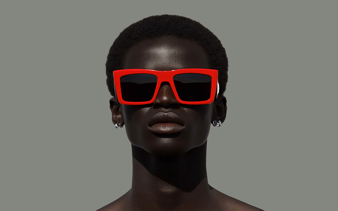Portrait of a person with short curly hair wearing large red sunglasses and silver hoop earrings against a plain gray background.