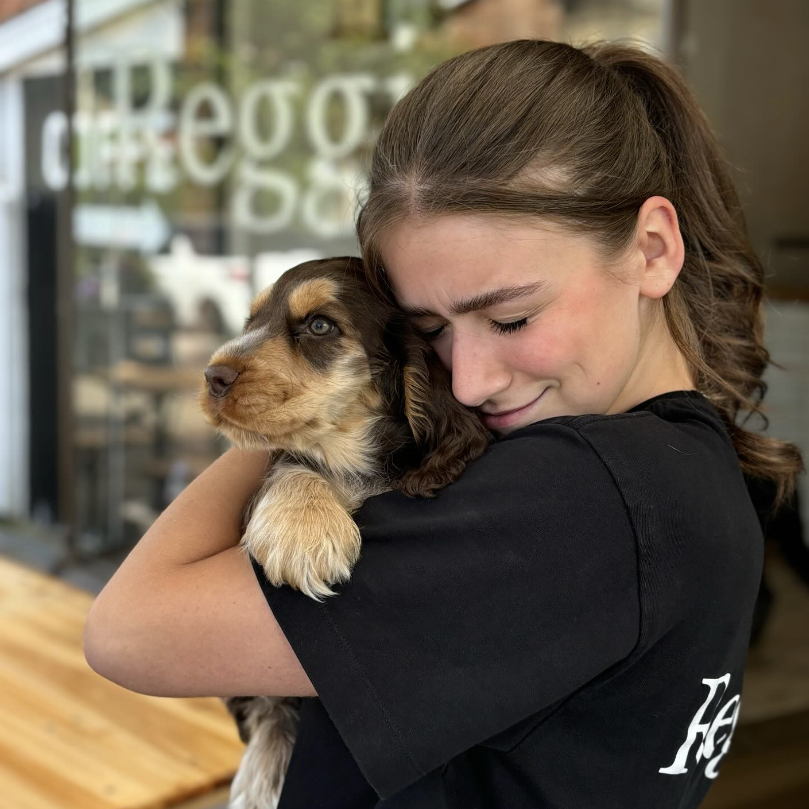 Young woman with brown hair hugging a brown and tan puppy closely with eyes closed and smiling.