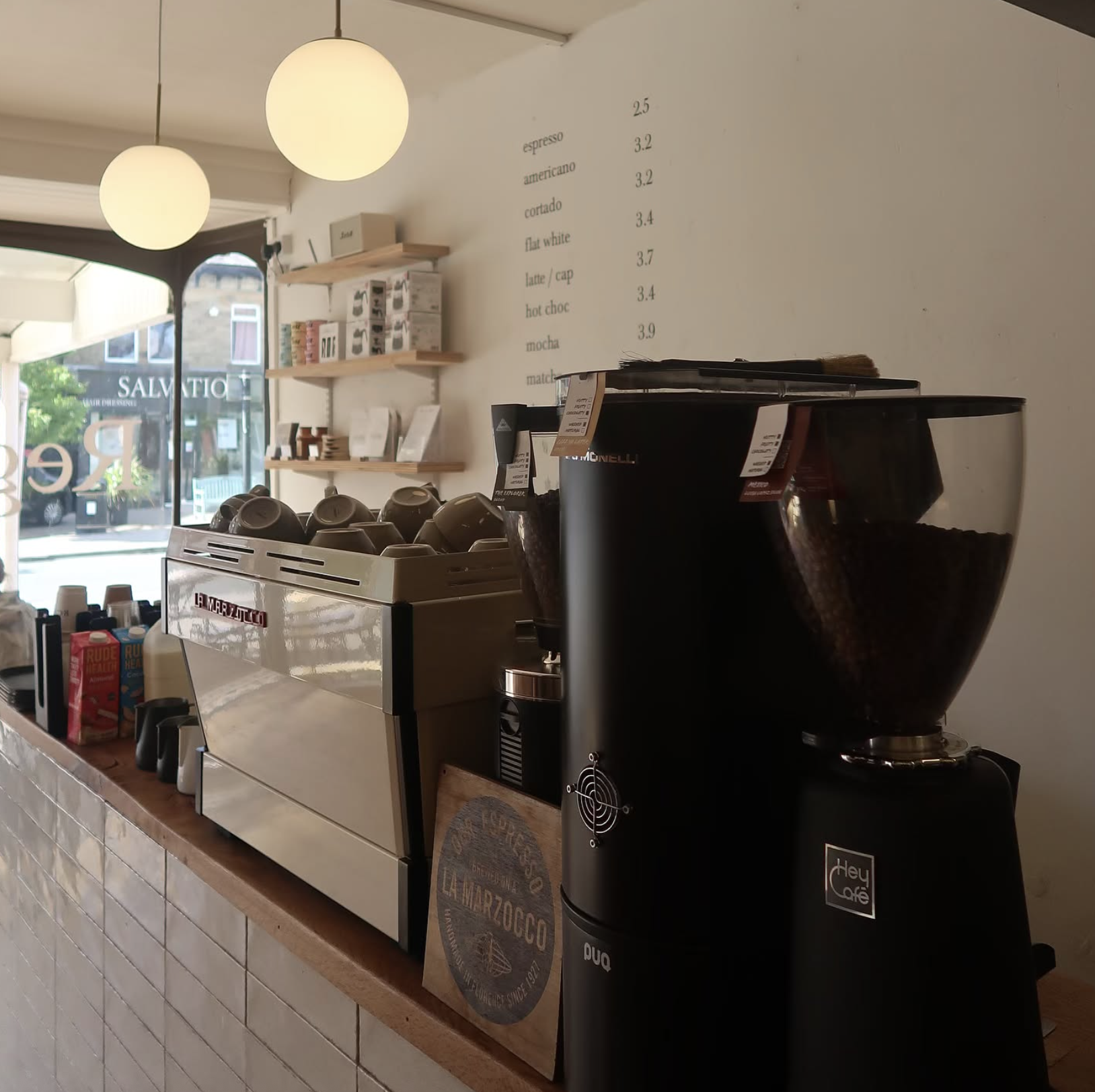 Counter in a coffee shop with a La Marzocco espresso machine, coffee grinders filled with beans, cups, and a menu on the wall.