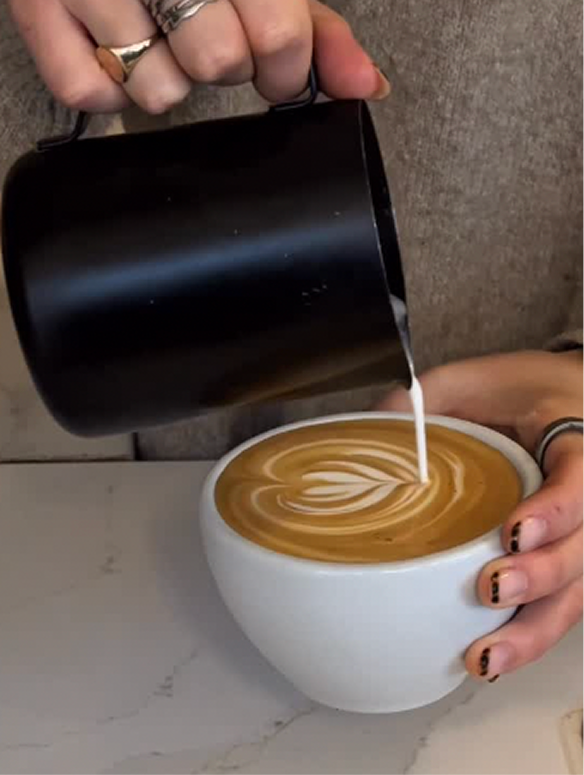 Hands with rings pouring steamed milk from a black pitcher into a cup of latte art coffee.