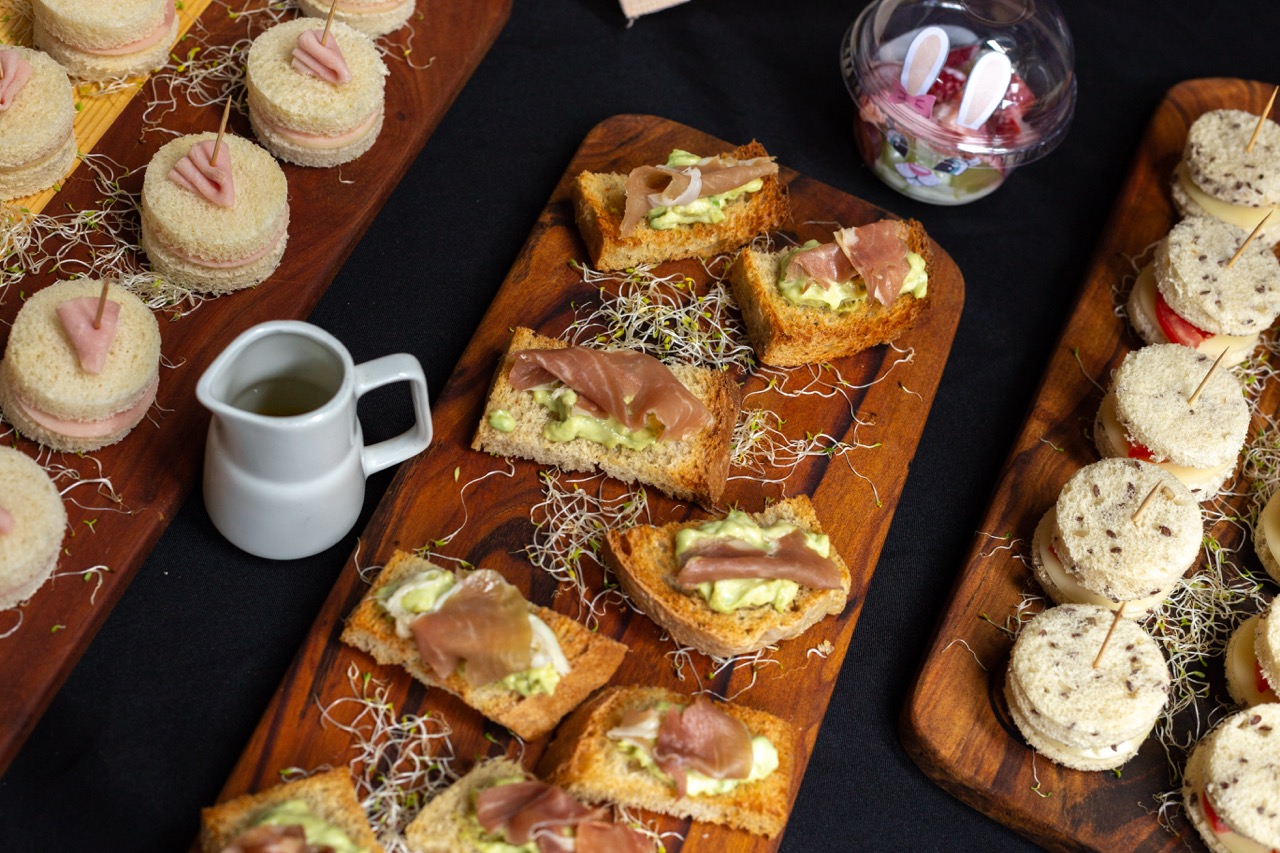 Wooden boards with assorted finger sandwiches topped with ham, avocado spread, and sprouts, alongside a small white pitcher.