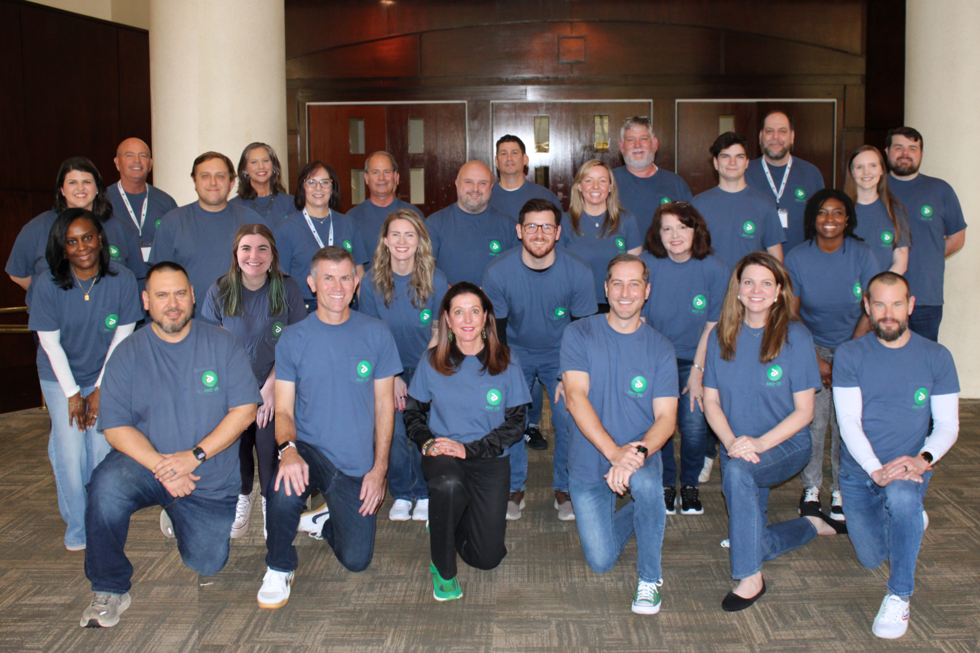 Group portrait of 27 people wearing matching blue t-shirts with a green logo, posed indoors in front of wooden doors.
