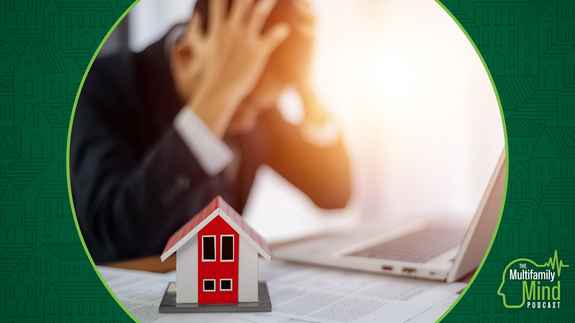 stressed businessman clutches head on desk in background with a model house in the foreground