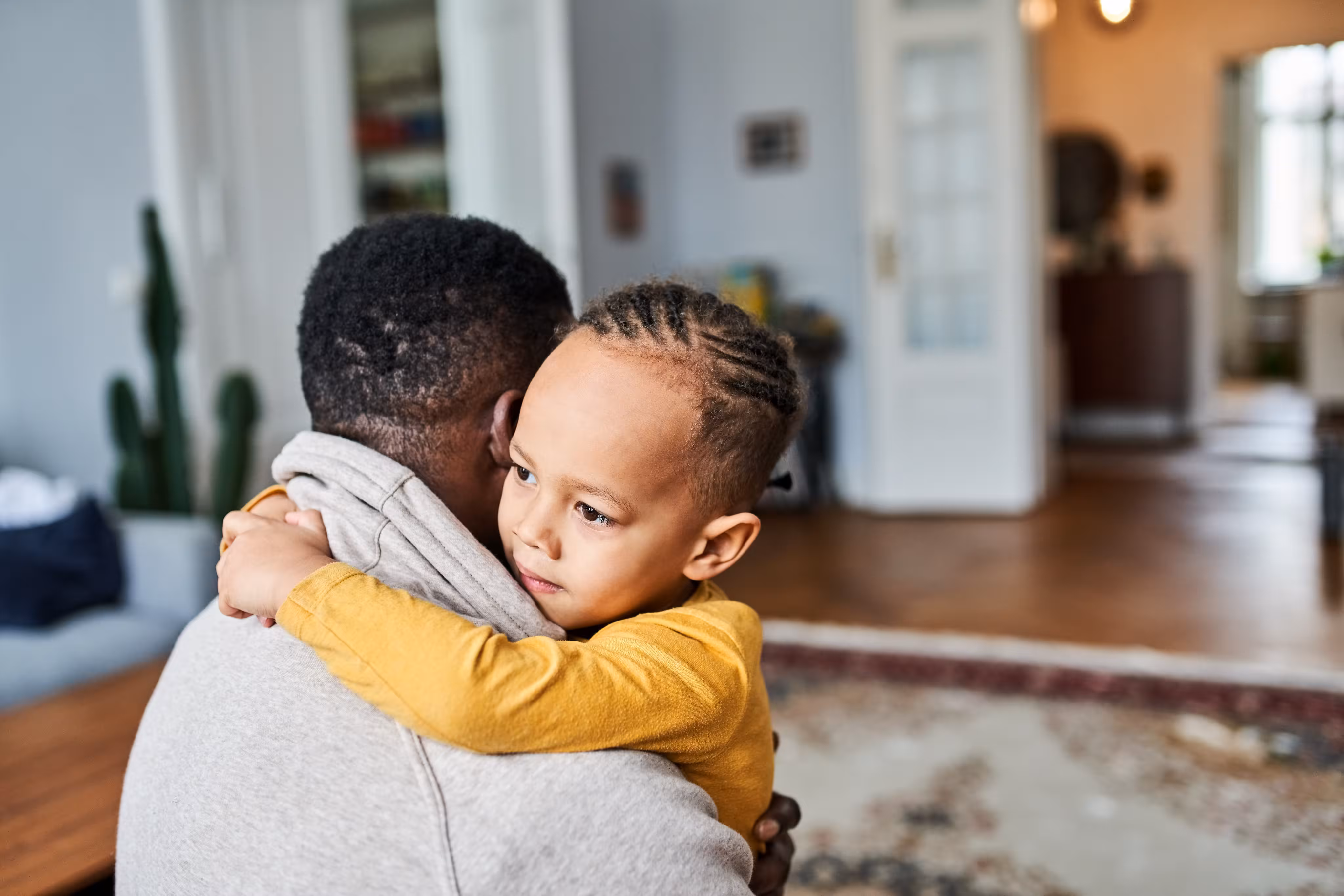 Young child with braids in a yellow shirt hugging an adult wearing a gray hoodie in a cozy living room.