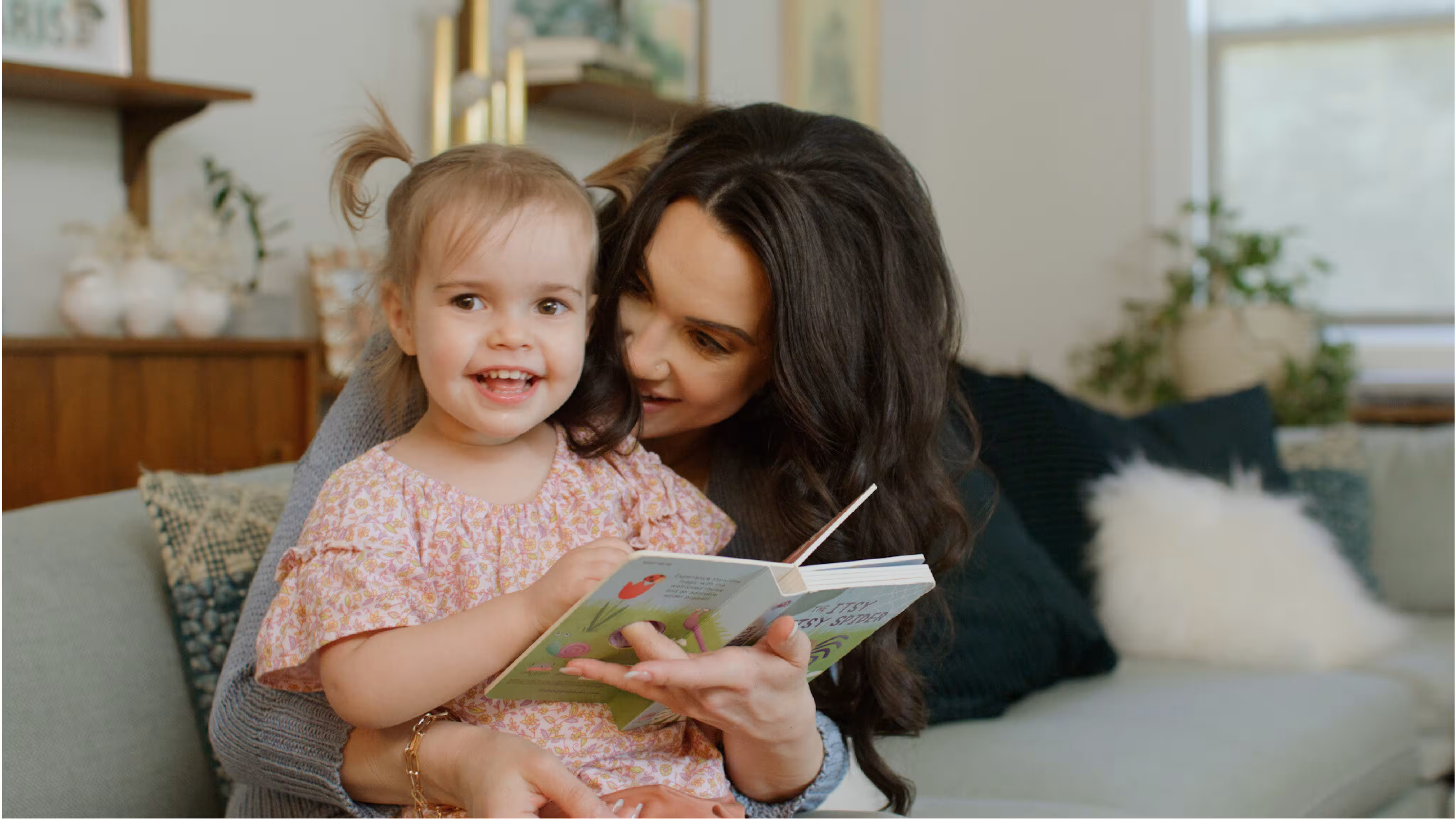 A woman with long dark hair reads a book to a smiling toddler girl in a floral dress while sitting on a sofa.