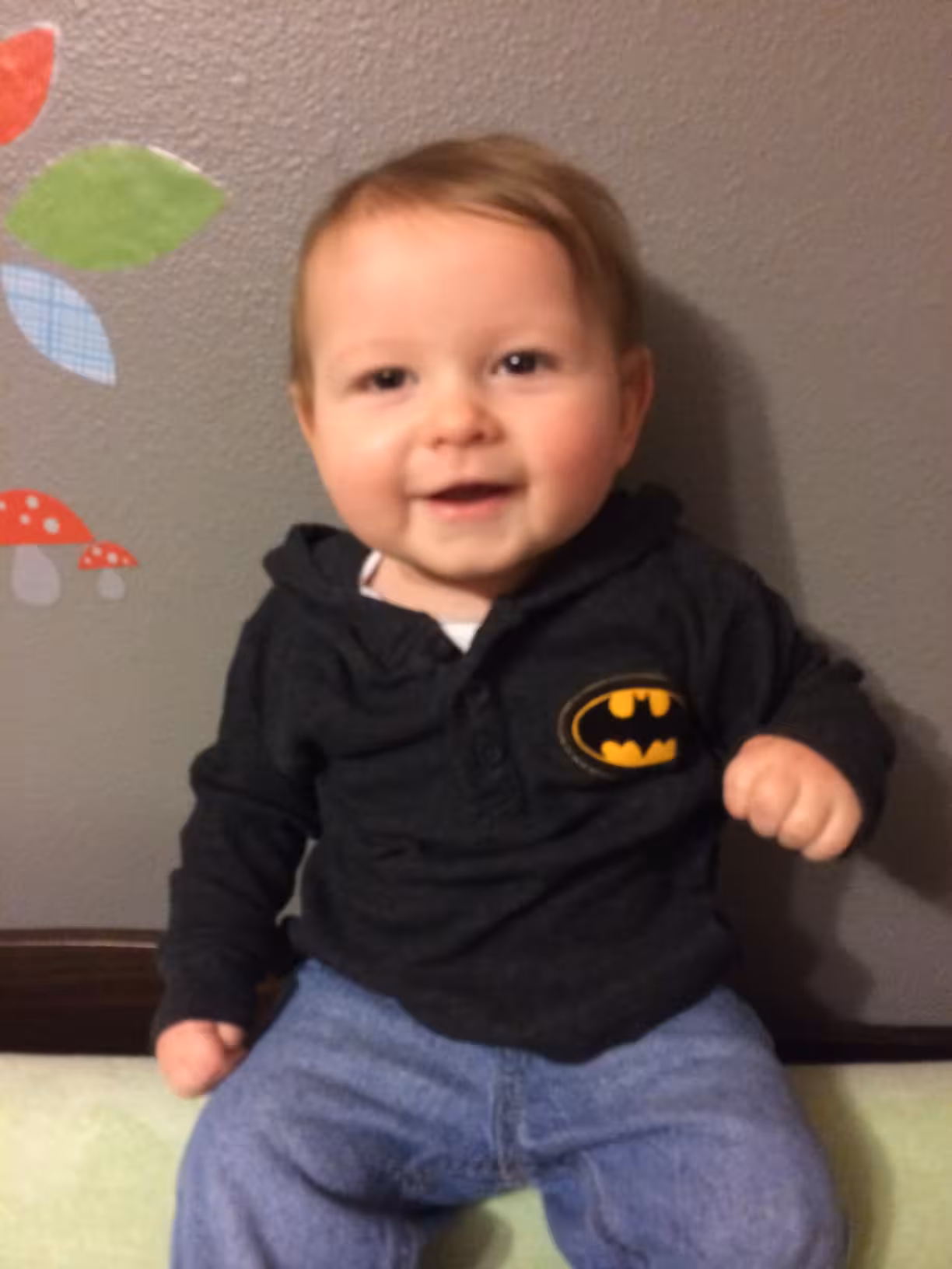 Smiling baby wearing a black Batman hoodie and blue jeans sitting against a gray wall.