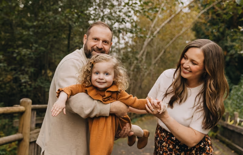Smiling parents holding and swinging their young daughter outdoors on a wooded path.