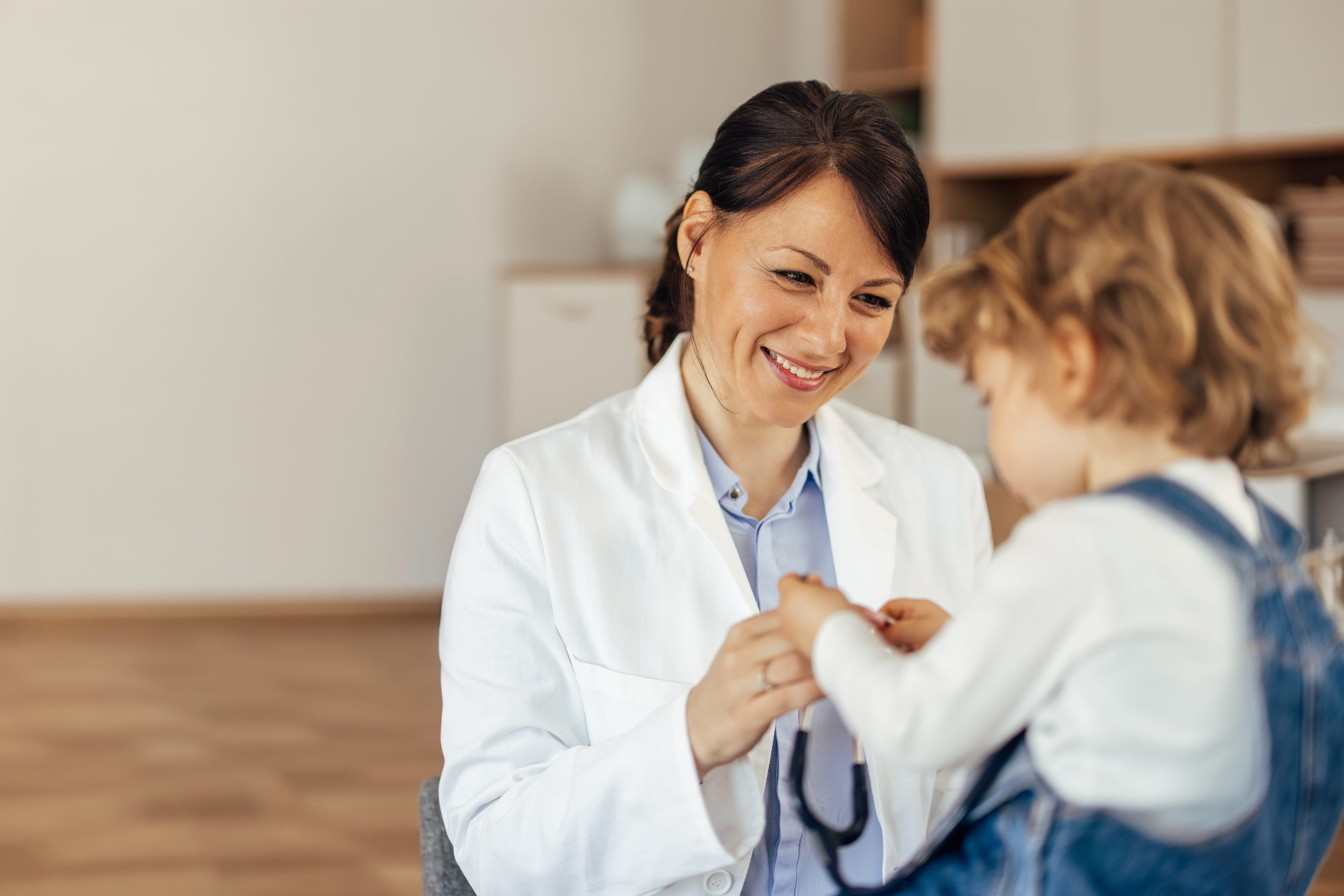 Smiling female doctor in white coat interacts with a toddler holding a stethoscope in a clinic.