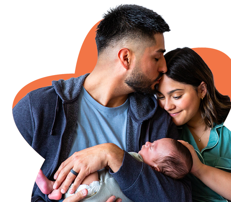 Man kisses woman on forehead while she smiles and they hold a sleeping newborn baby together.