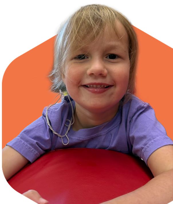 Smiling young child with blonde hair wearing a purple shirt leaning on a red surface.