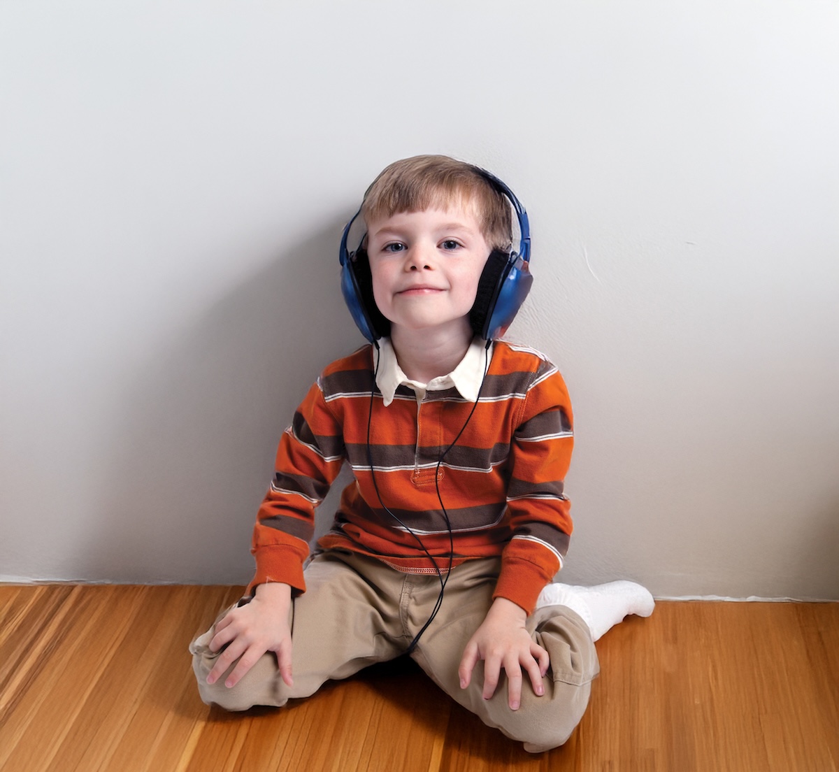 Young boy with headphones sitting cross-legged on wooden floor against white wall.