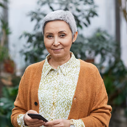 Smiling mature woman with short gray hair holding a smartphone, wearing a patterned shirt and brown cardigan.