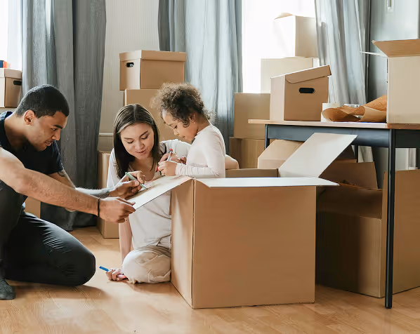 Family with a young child packing or unpacking cardboard boxes in a room with hardwood floors.