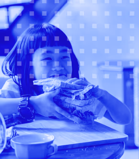 Child holding an oversized sandwich with both hands at a table with a cup nearby.