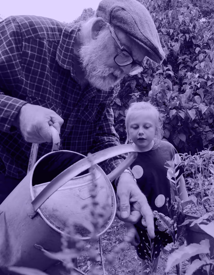 Older man watering plants while a young girl watches and points at a plant in a garden.