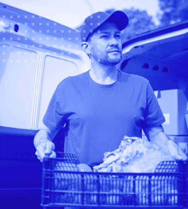 Man wearing cap holding a crate of groceries outside a van under a partly cloudy sky.