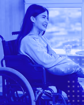 Smiling woman sitting in a wheelchair indoors near a window holding a pillow on her lap.