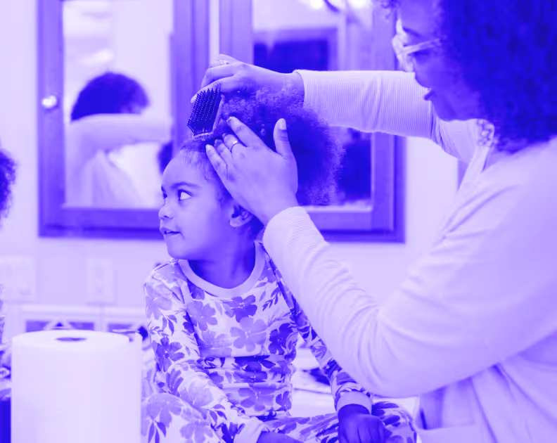 Adult combing a young girl's curly hair as she sits and looks to the left in a bathroom.