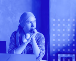 Woman wearing headscarf sitting and looking thoughtfully out a window with hands clasped.