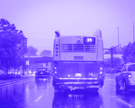 Rear view of a city bus on a rainy street with cars and trees around during dusk.