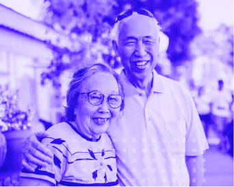 Smiling elderly couple outdoors, man with arm around woman, both wearing glasses and casual clothes.