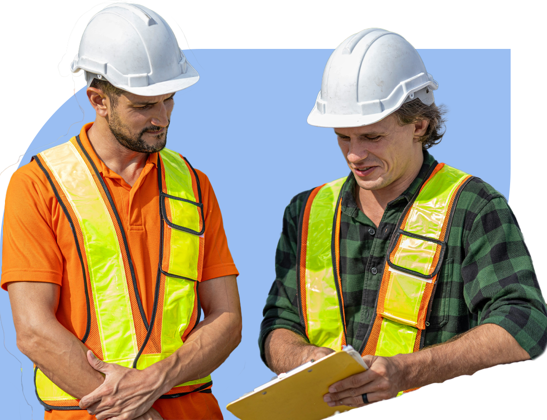Dos trabajadores de construcción con cascos blancos y chalecos reflectantes revisan un portapapeles; uno sostiene el documento mientras ambos lo observan con atención.