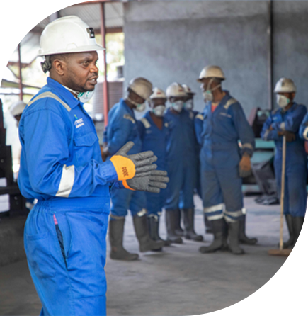 Trabajador industrial portando un uniforme azul, casco protector blanco, y guantes, frente a diferentes trabajadores ubicados al fondo de la imagen que portan el mismo uniforme.
