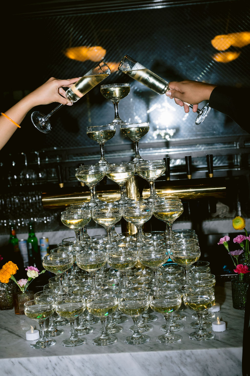 Two hands pouring champagne over a tower of coupe glasses