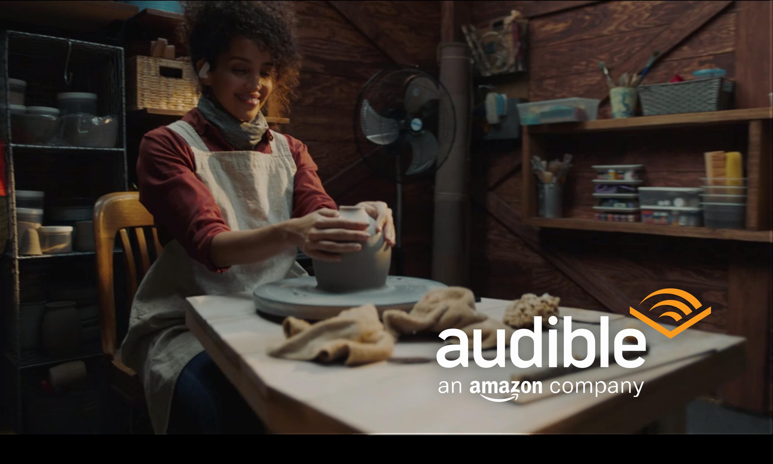 Smiling woman wearing an apron shaping clay on a pottery wheel in a wooden workshop with the Audible logo and text 'an Amazon company' in the lower right corner.