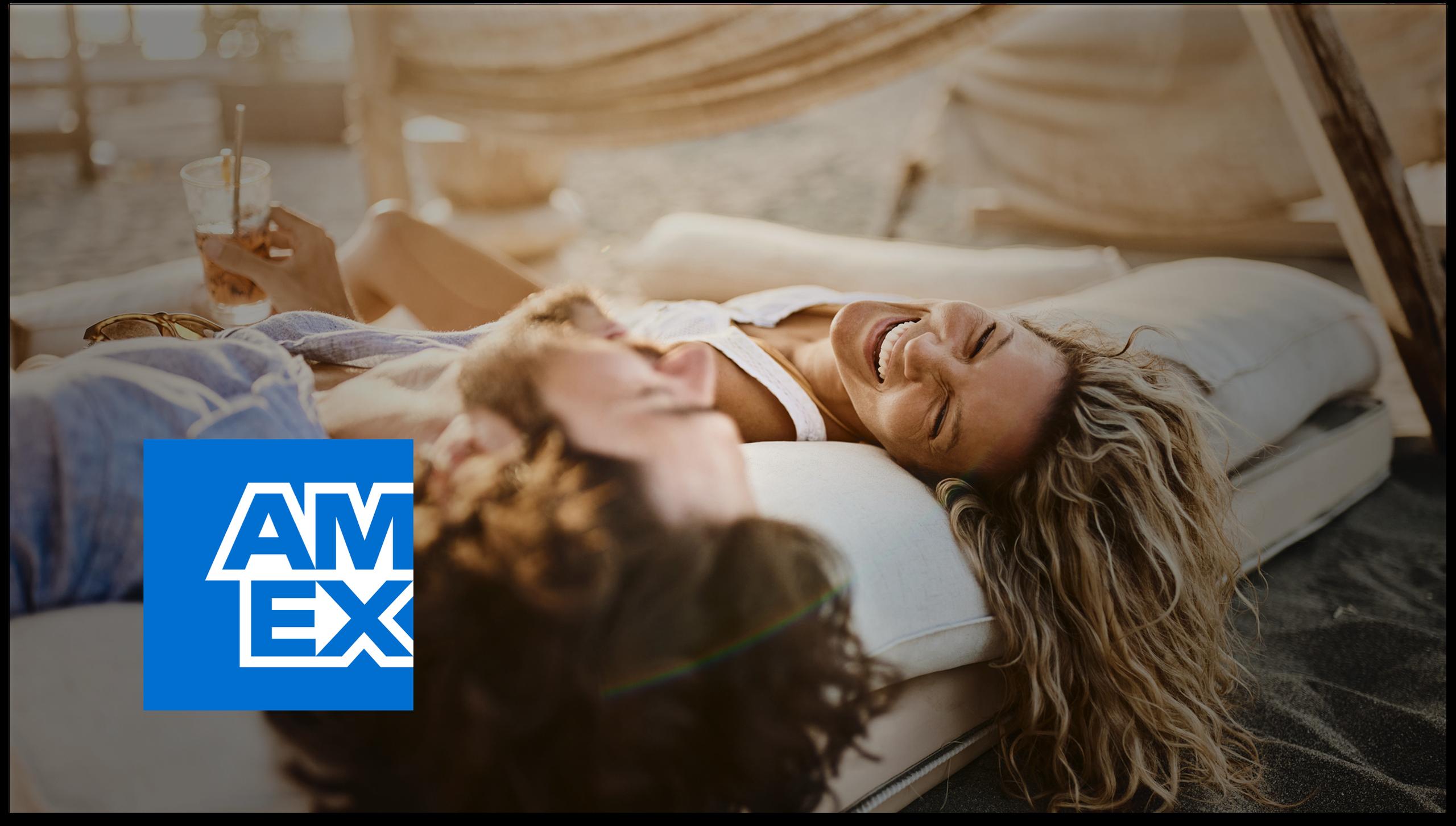 Smiling couple lying on cushions at the beach with an AMEX logo in the corner.