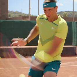 Tennis player wearing a green shirt and blue shorts hitting a forehand shot on a clay court.