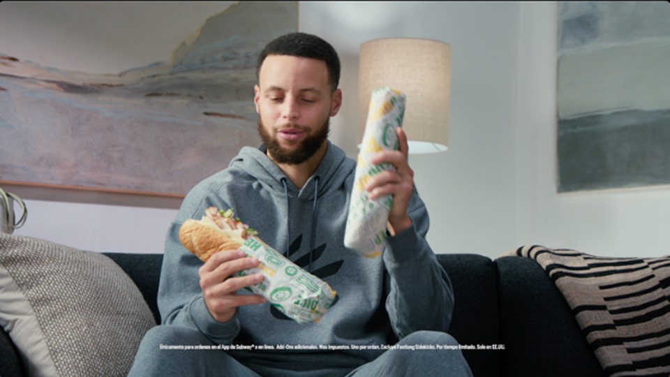 Man in a gray hoodie sitting on a couch holding two wrapped Subway sandwiches, with a floor lamp and abstract paintings in the background.