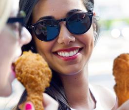 Close-up of two women smiling and holding fried chicken drumsticks, one woman wearing sunglasses.
