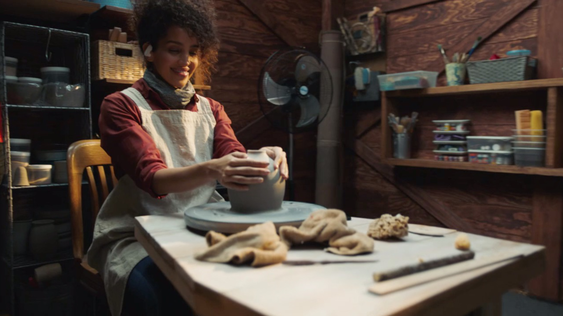 Smiling woman shaping clay on a pottery wheel in a wood-paneled workshop.