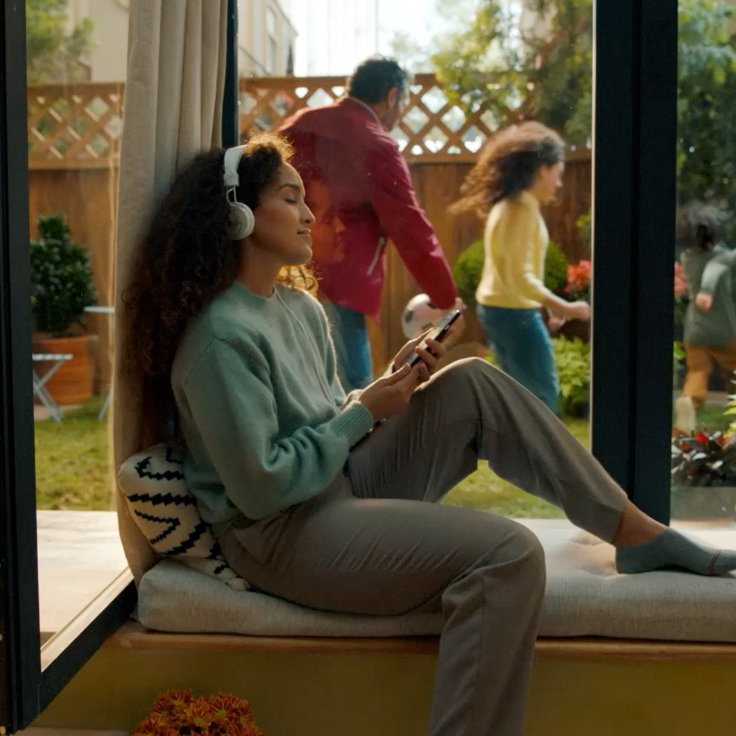 Young woman with headphones sitting by a window, holding a smartphone, while a man and two children play outside in a garden.
