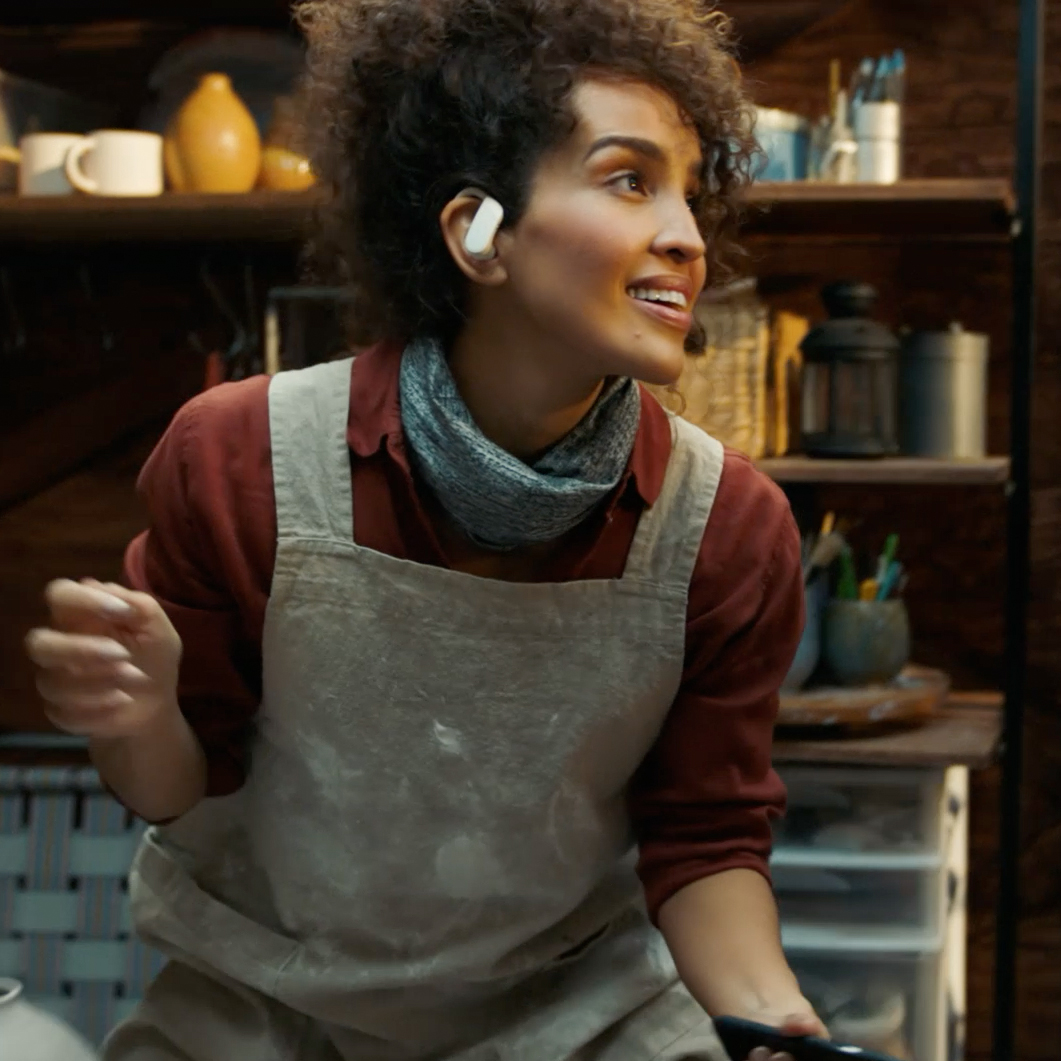 Smiling woman with curly hair wearing a beige apron and wireless earbuds in a cozy workshop.