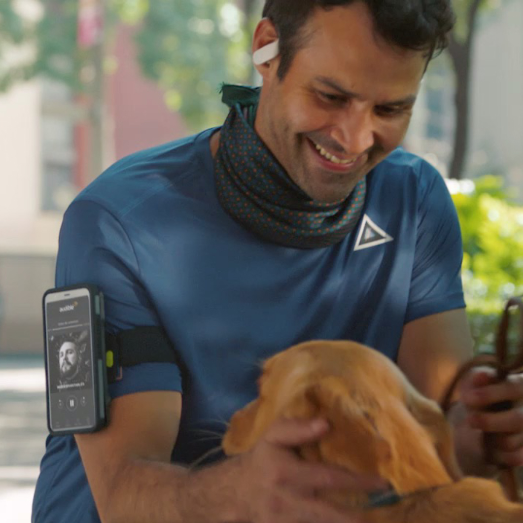 Smiling man in blue workout shirt with a phone on his arm and earbuds, petting a golden retriever outdoors.