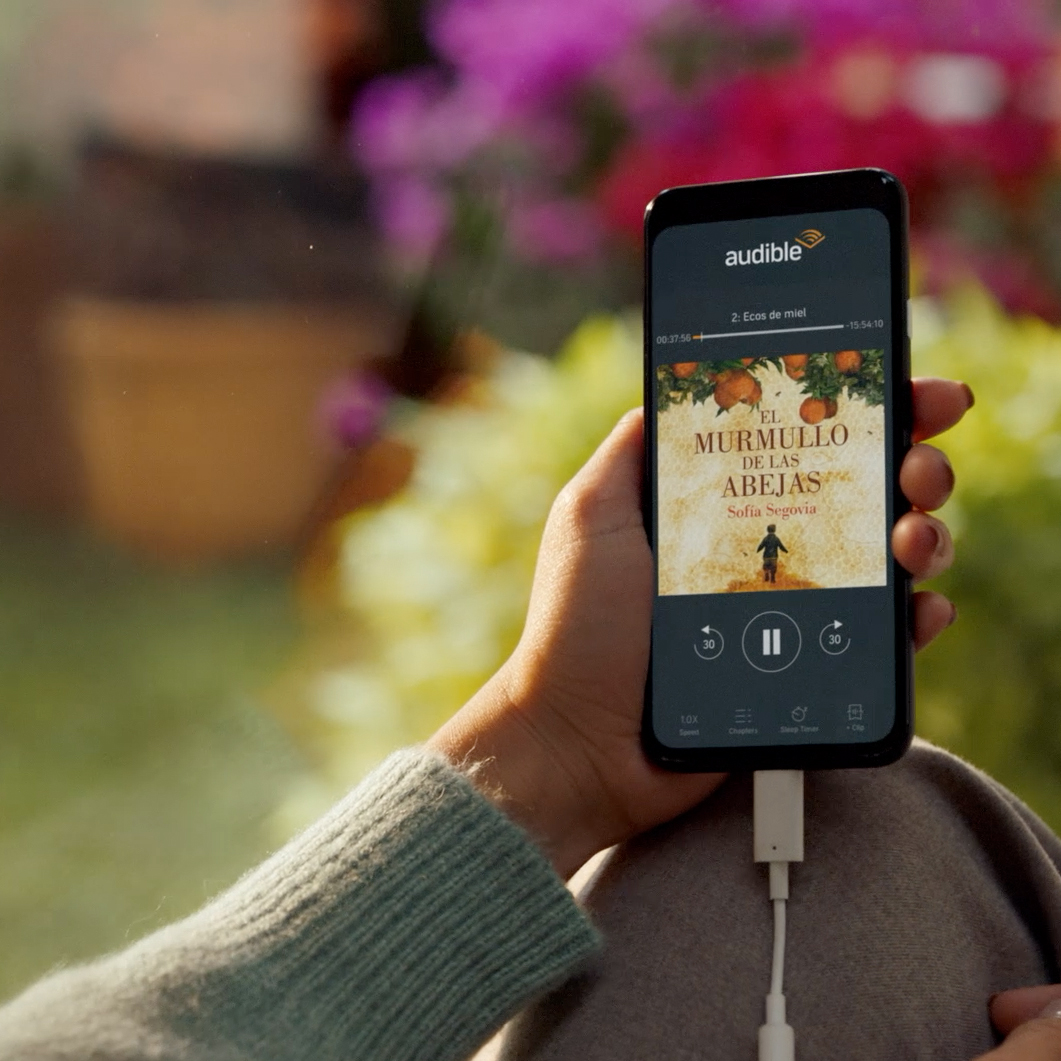 Hand holding a phone playing the Audible audiobook 'El Murmullo de las Abejas' by Sofía Segovia with colorful flowers blurred in the background.