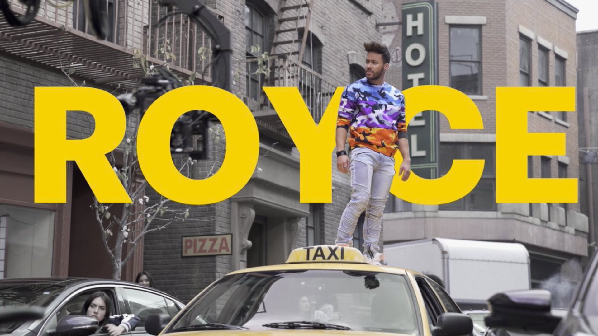 Man wearing a colorful camo sweater and ripped jeans standing on top of a yellow taxi in a city street with large yellow text reading ROYCE behind him.
