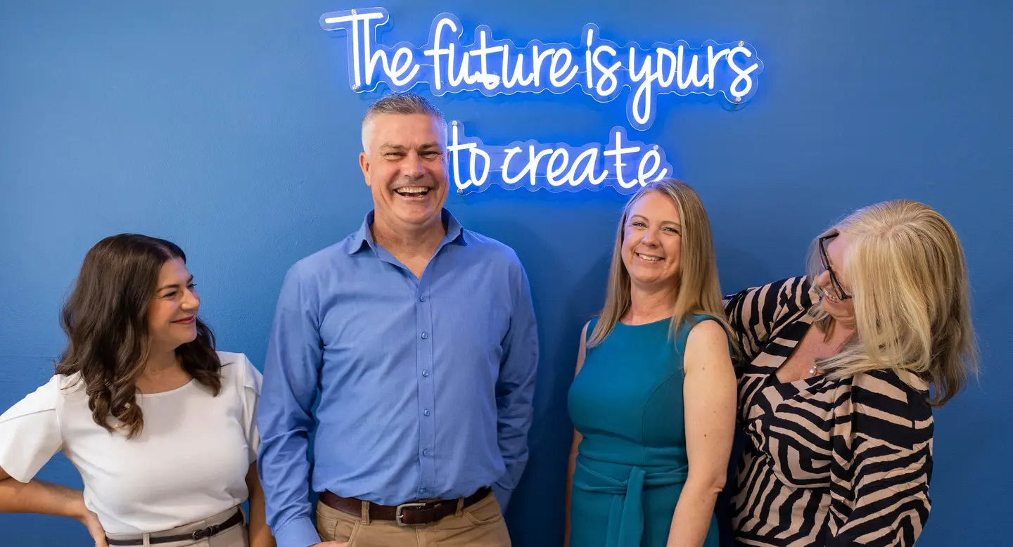 Four smiling people posing in front of a blue wall with a neon sign that reads 'The future is yours to create.'