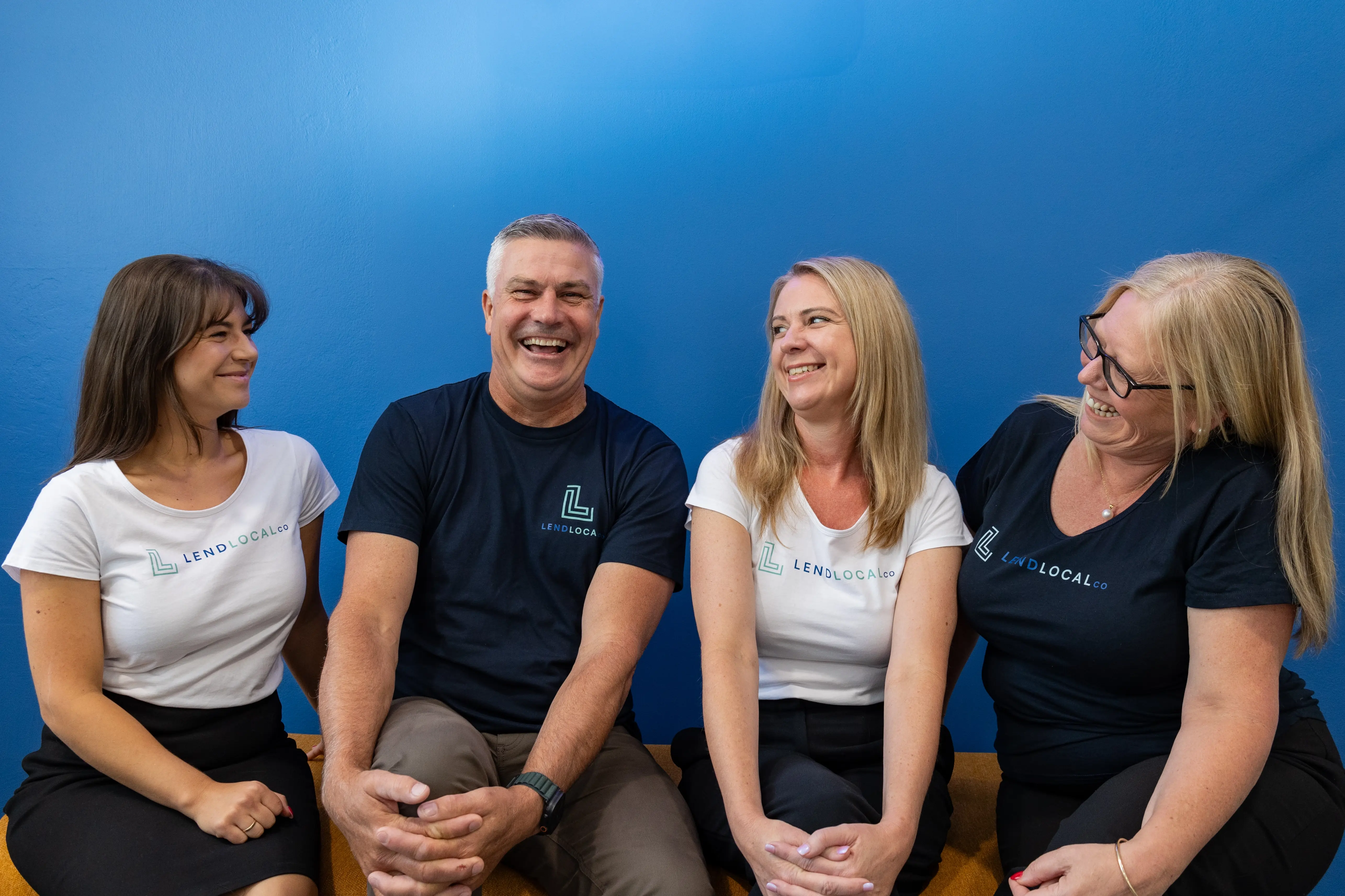 Four people sitting on a couch in front of a blue wall, smiling and wearing black and white LendLocal branded t-shirts.