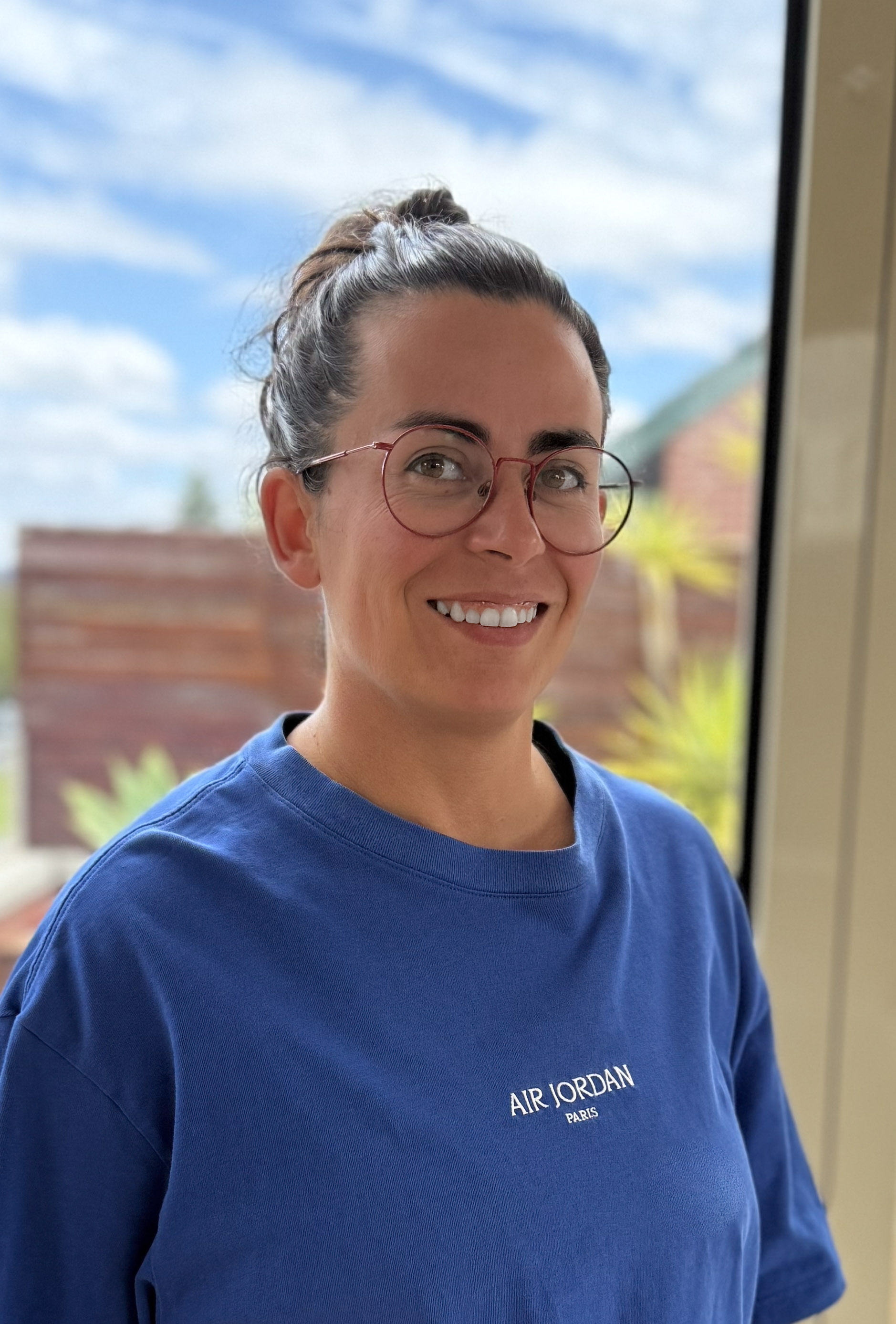 Smiling woman with glasses and hair tied back wearing a blue Air Jordan Paris T-shirt indoors near a window.